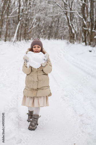 a cute six-year-old girl stands in a winter forest and holds a snowball