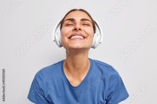 Photo of pleased satisfied woman keeps eyes closed from pleasure catches every bit of song listens music via wireless headphones smiles toothily wears casual blue t shirt poses against white wall