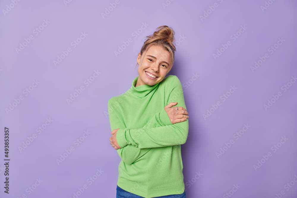 Pleased lovely woman with combed hair embraces herself feels comfortable wears green jumper smiles gently being self egoist poses against purple background. Self love and acceptance concept. Pleased lovely woman with combed hair embraces herself feels comfortable wears green jumper smiles gently being self egoist poses against purple background. Self love and acceptance concept.
