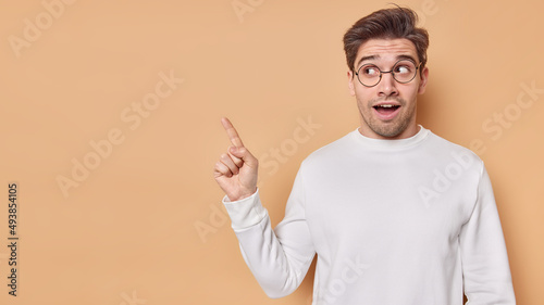 Horizontal shot of surprised handsome young man points at copy space shows awesome promotion or advertisement has amazed facial expression wears casual white jumper spectacles. Wow look at this