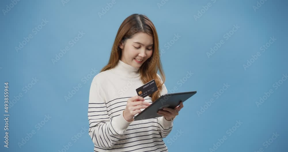 Youth cheerful Asia lady wear casual sweater internet shopping woman online with digital tablet and credit card stand isolated over blue background. Female use debit card payment online banking. Youth cheerful Asia lady wear casual sweater internet shopping woman online with digital tablet and credit card stand isolated over blue background. Female use debit card payment online banking.