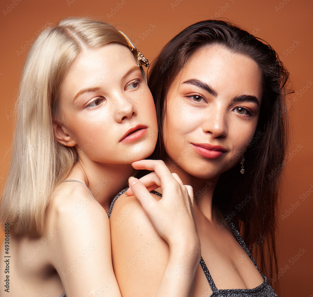 two pretty diverse girls happy posing together: blond and brunette, caucasian and asian on brown background, lifestyle people concept