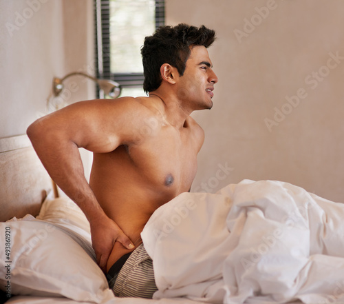 Bad quality sleep leaves him feeling stiff and sore. Shot of a young man waking up in bed with back pain.
