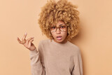 Unhappy young curly haired woman feels indignant keeps hand raised feels clueless dressed in casual jumper isolated over beige studio background cannot understand something. So what did you say
