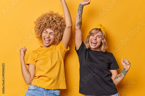 Slika na platnu Horizontal shot of happy young beautiful women enjoy life feel carefree shake arms cheer over something chill indoor dressed in casual clothes isolated over yellow background