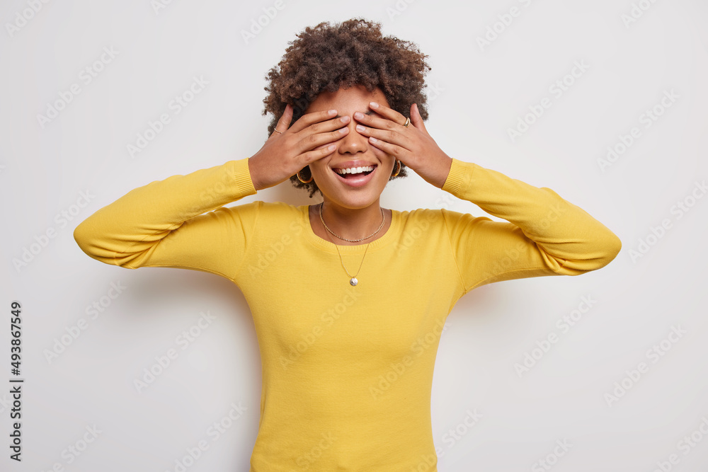 Positive curly haired young woman plays hide and seek covers eyes with palms smiles waits for surprise wears yellow jumper isolated over white background. Glad female model makes blind gesture Positive curly haired young woman plays hide and seek covers eyes with palms smiles waits for surprise wears yellow jumper isolated over white background. Glad female model makes blind gesture
