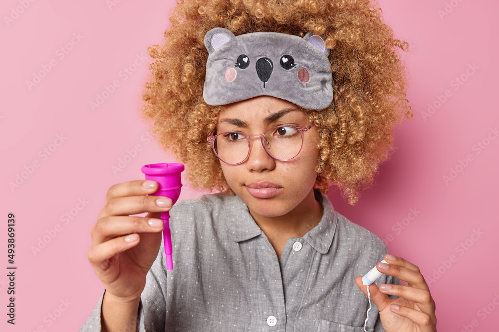 Serious curly haired young woman examines new product for menses holds menstruation cup and clean tampon has attentive gaze wears sleepmask and pajama isolated over pink background. Periods concept