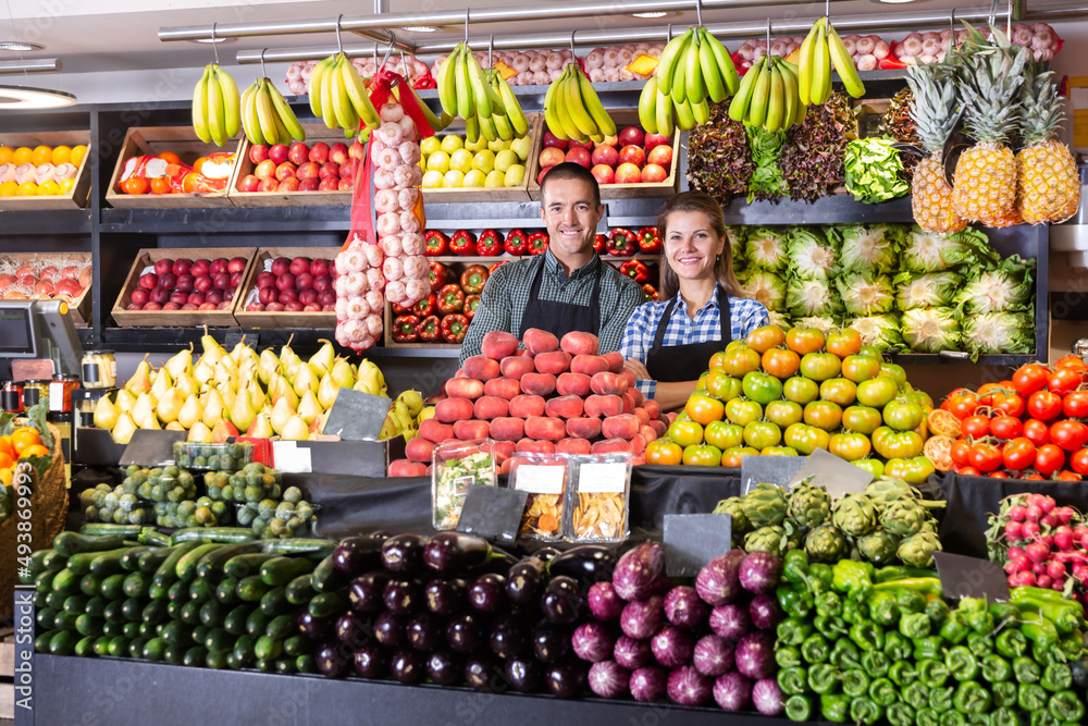 Young man and woman sellers in aprons standing near counter with fresh vegetables and fruits at market