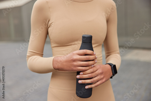 Cropped image of unknown sportswoman dressed in beige activewear holds bottle of fresh cold refreshing water uses smartwatch poses outdoors against blyurred background. Healthy lifestyle concept