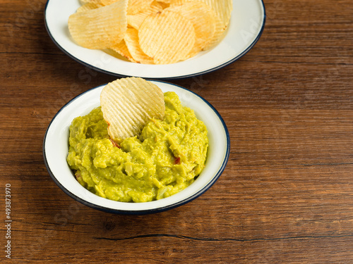 A bowl with fresh guacamole and potatoes chips on a white plate over a wooden table