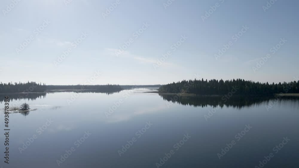 Drone flying slowly close to a large calm lake surround by green forest under a blue sky. Reflection can be seen in the water. Drone flying slowly close to a large calm lake surround by green forest under a blue sky. Reflection can be seen in the water.