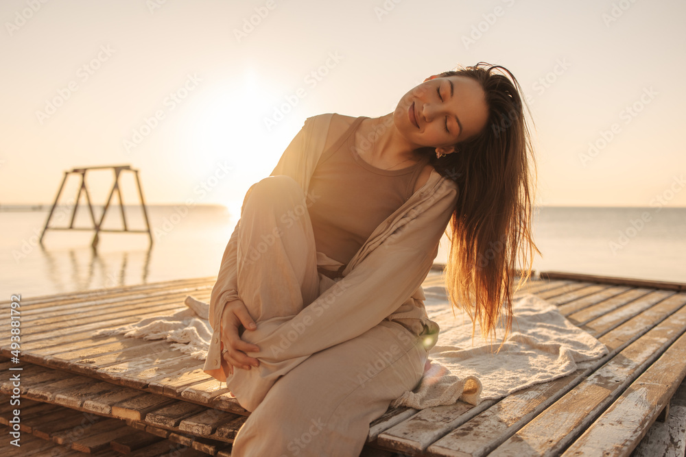 Relaxed young fair-skinned girl sits on pier with closed eyes on ocean shore. Model has leg pressed to her, head tilted to side. Concept of rest and recovery.