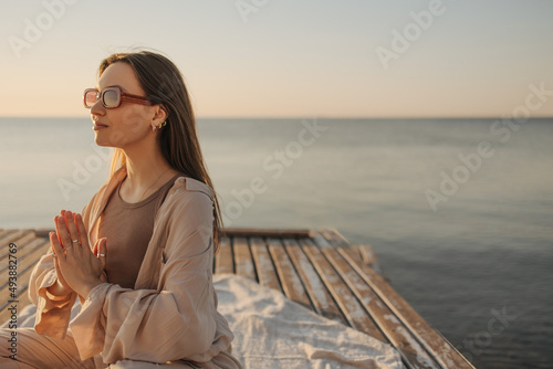 Close-up of young caucasian woman holding palms pressed together, dressed casual outfit on seashore. Girl in sunglasses sits on pier and shows prayer gesture, hoping that dreams will come true.