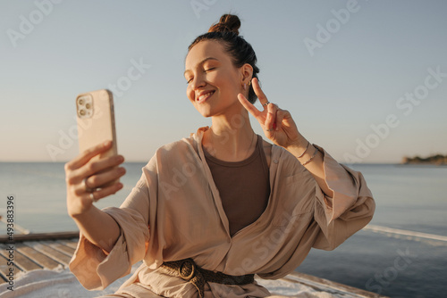 Happy fair-skinned young lady is photographed on modern smartphone showing two fingers. Blogger girl broadcasts video with her eyes closed on seashore. Happy weekend concept.