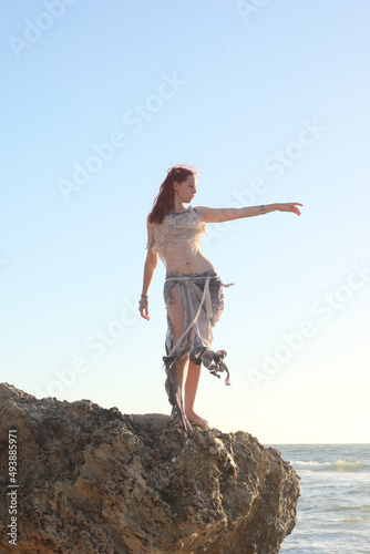 Full length portrait of red haired woman wearing torn shipwrecked clothing. Standing pose with gestural hands at rocky ocean beach landscape background. 