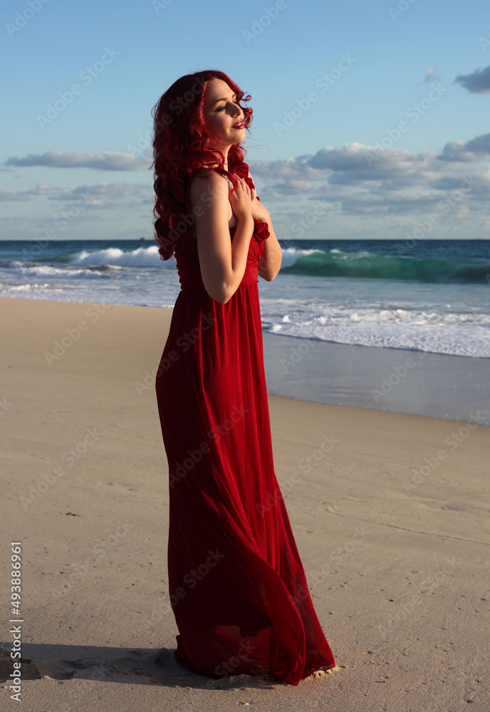 Full length portrait of redhead woman wearing elegant gown. Standing pose with gestural hands at sunset ocean beach landscape background. Full length portrait of redhead woman wearing elegant gown. Standing pose with gestural hands at sunset ocean beach landscape background.