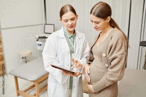 Waist up portrait of young female doctor consulting pregnant woman in clinic and using digital tablet