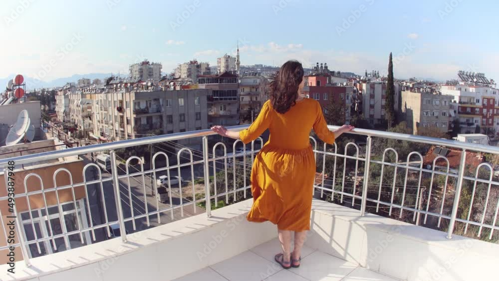 A girl in a yellow dress stands on a balcony during a strong wind, a smiling woman stands on a roof overlooking the city and the wind inflates her dress, an attractive girl looks at the city