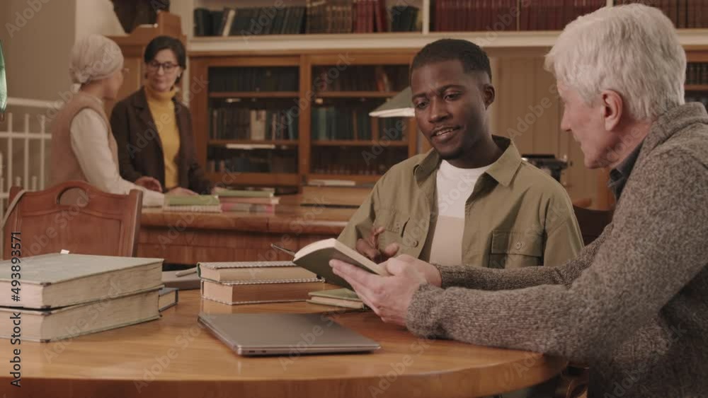 Medium slowmo of African American male university student and his senior professor of Literature discussing book plot while studying in library with two women talking in background Medium slowmo of African American male university student and his senior professor of Literature discussing book plot while studying in library with two women talking in background