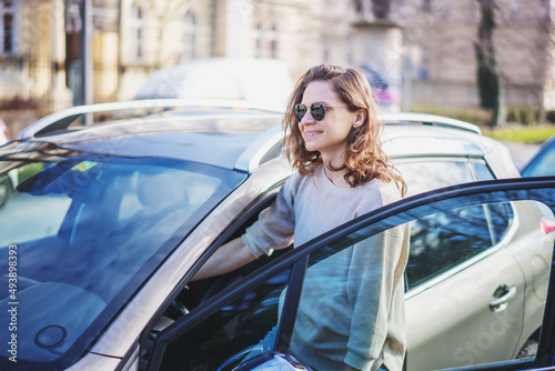 Happy cheerful young woman in sunglasses getting into her car on a city street