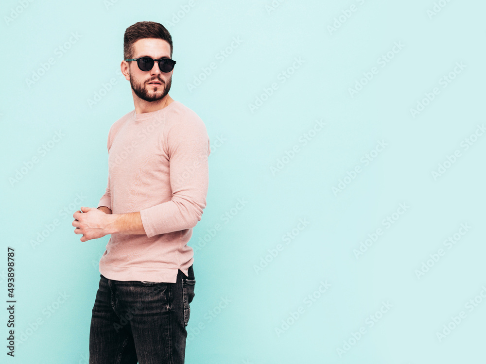 Portrait of handsome confident model. Sexy stylish man dressed in pink sweater and jeans. Fashion hipster male posing near blue wall in studio. In sunglasses. Isolated Portrait of handsome confident model. Sexy stylish man dressed in pink sweater and jeans. Fashion hipster male posing near blue wall in studio. In sunglasses. Isolated