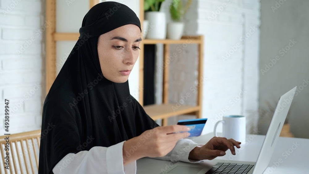 A woman in a hijab makes an online payment using a credit card and a laptop. Remote payment in the Islamic world. When buying in an online store, a Muslim woman enters her card details for payment. A woman in a hijab makes an online payment using a credit card and a laptop. Remote payment in the Islamic world. When buying in an online store, a Muslim woman enters her card details for payment.