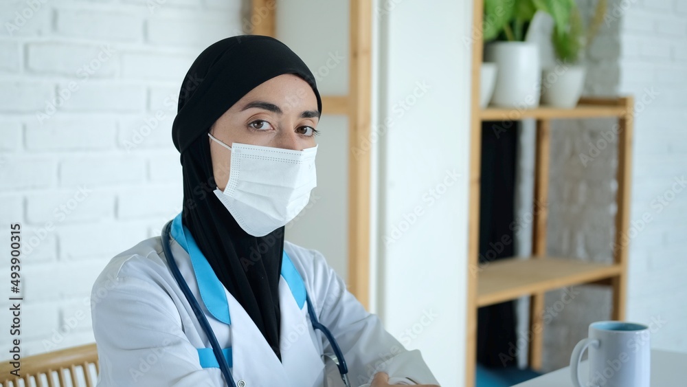 Muslim woman doctor in a mask looks at the camera. Working in a hospital for women in the Arab world. A medical worker in a black veil and medical gown at the workplace.