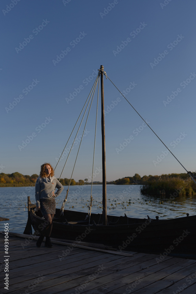 A laughing young redhead woman enjoying a spring day at the lake, standing on a wooden pier above a boat