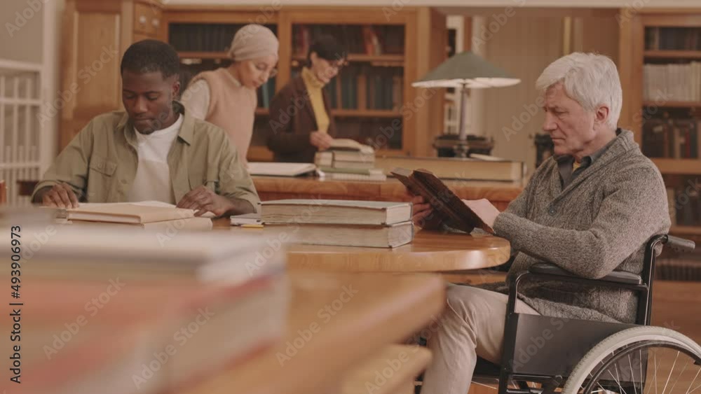 Medium slowmo of senior Caucasian man with disability reading book sitting in wheelchair at reading room of big library with other people studying or reading quietly Medium slowmo of senior Caucasian man with disability reading book sitting in wheelchair at reading room of big library with other people studying or reading quietly
