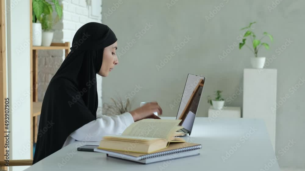 A Muslim female teacher reads a book and types learning material on a laptop keyboard. The rights of Muslim women to education. A woman in a black hijab conducts remote training. A Muslim female teacher reads a book and types learning material on a laptop keyboard. The rights of Muslim women to education. A woman in a black hijab conducts remote training.