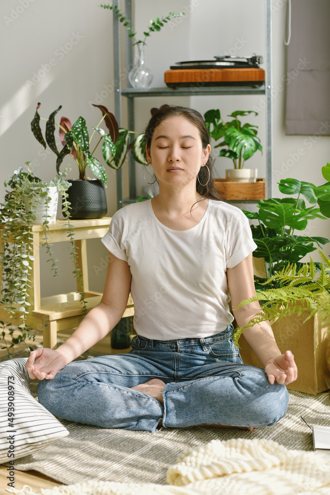 Asian woman practice meditation in lotus position for relieve stress and tension, Woman sitting in living room decor with green houseplant .