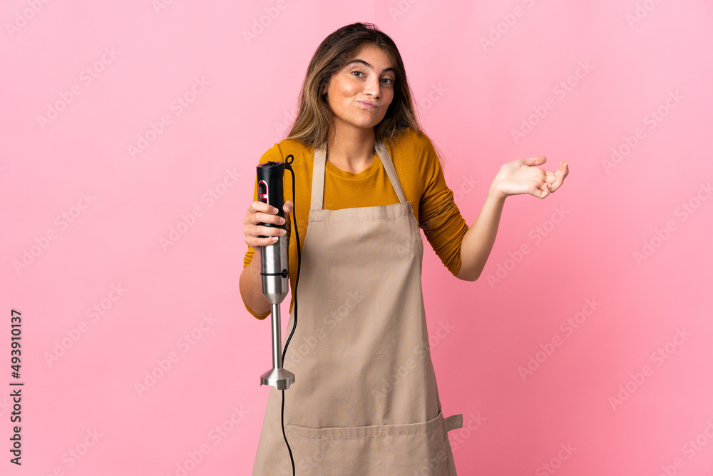 Young chef woman using hand blender isolated on pink background having doubts while raising hands Young chef woman using hand blender isolated on pink background having doubts while raising hands