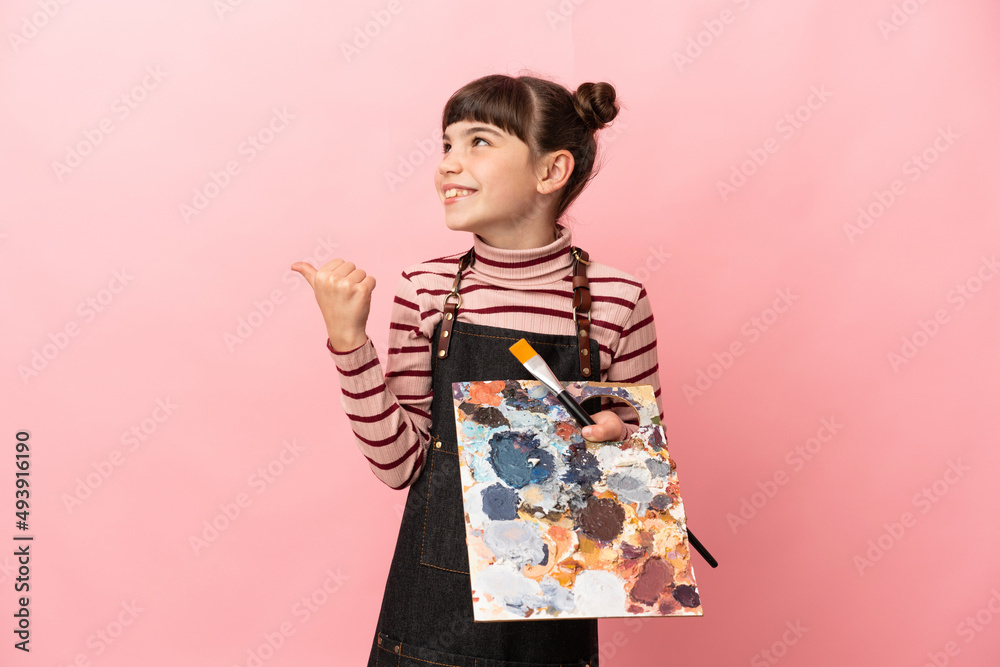 Little artist girl holding a palette isolated on pink background pointing to the side to present a product Little artist girl holding a palette isolated on pink background pointing to the side to present a product