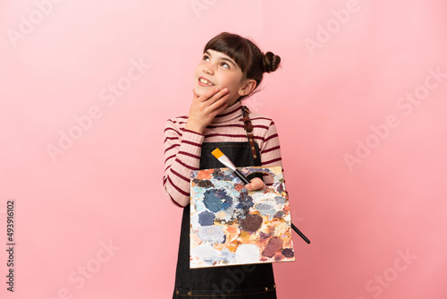 Little artist girl holding a palette isolated on pink background looking up while smiling