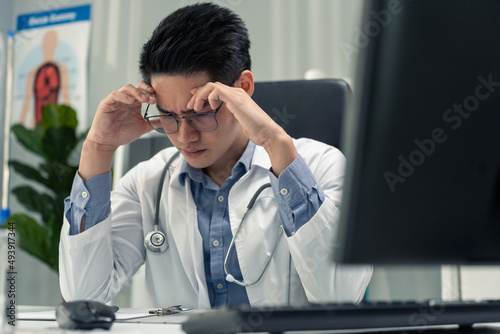 Stressed and overworked Asian male doctor sitting on table in hospital. 