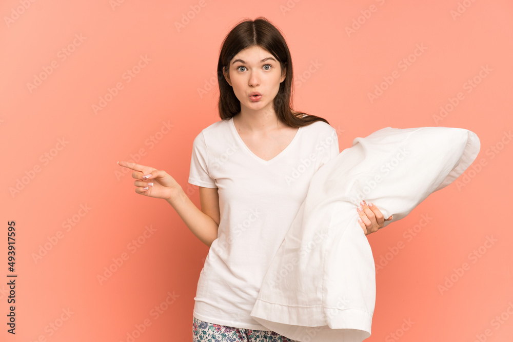 Young Ukrainian girl isolated on pink background in pajamas and surprised while pointing side Young Ukrainian girl isolated on pink background in pajamas and surprised while pointing side