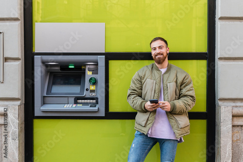 Smiling young man with smart phone standing by atm photo