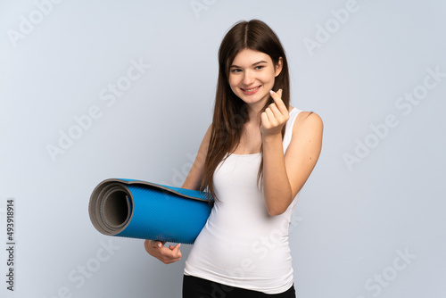 Young Ukrainian sport girl going to yoga classes while holding a mat making money gesture