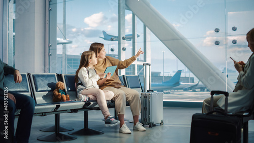 Busy Airport Terminal: Cute Mother and Little Daughter Wait for their Vacation Flight, Play Educational Games on Digital Tablet. People Sitting in Boarding Lounge of Airline Hub with Airplanes Flying
