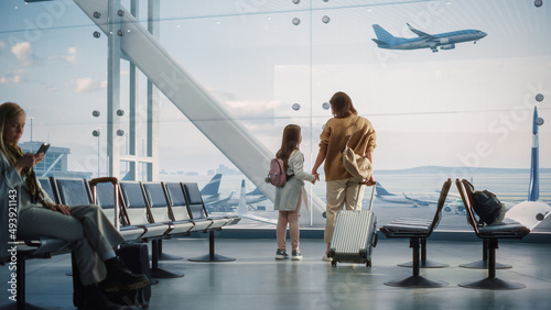 Airport Terminal: Beautiful Mother and Cute Little Daughter Wait for their Vacation Flight, Looking out of Window for Arriving and Departing Airplanes. Young Family in Boarding Lounge of Airline Hub photo