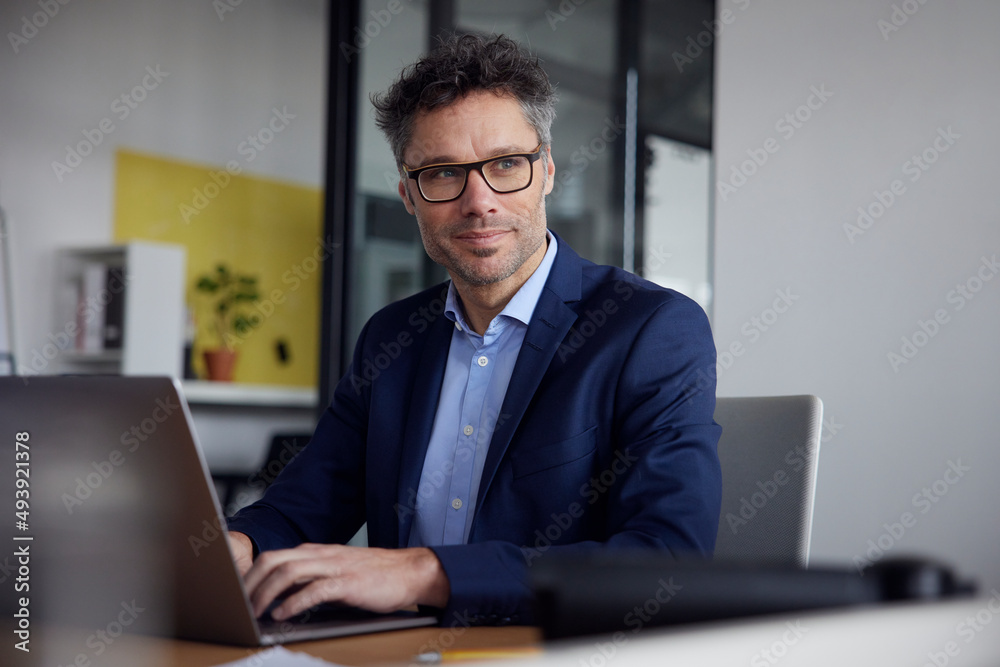 Thoughtful businessman wearing eyeglasses sitting with laptop at desk in office Thoughtful businessman wearing eyeglasses sitting with laptop at desk in office