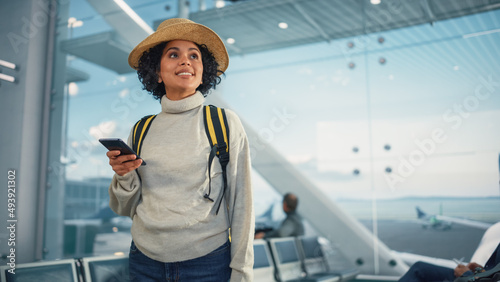Airport Terminal: Happy Traveling Black Woman Waiting at Flight Gates for Plane Boarding, Uses Mobile Smartphone, Checking Trip Destination on Internet. Smiling African American Female on Vacation photo