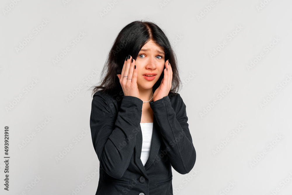 Portrait of startled young brunette woman grabbing head with both hands in despair , staring shocked and confused at camera, standing in black blazer over grey background Portrait of startled young brunette woman grabbing head with both hands in despair , staring shocked and confused at camera, standing in black blazer over grey background
