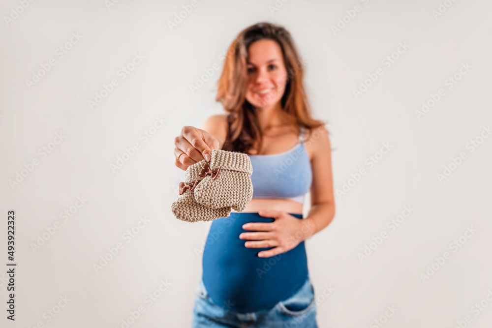 A happy pregnant woman in special jeans holds baby booties against a light wall. A woman in disfocus A happy pregnant woman in special jeans holds baby booties against a light wall. A woman in disfocus
