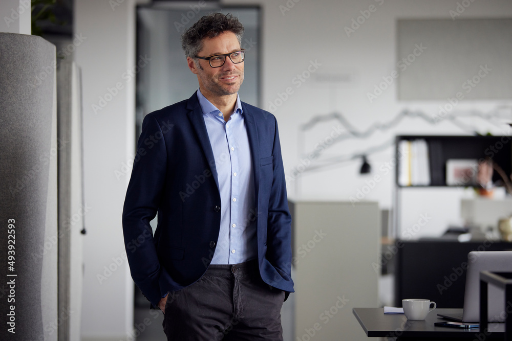 Smiling businessman wearing eyeglasses standing with hands in pockets at work place Smiling businessman wearing eyeglasses standing with hands in pockets at work place