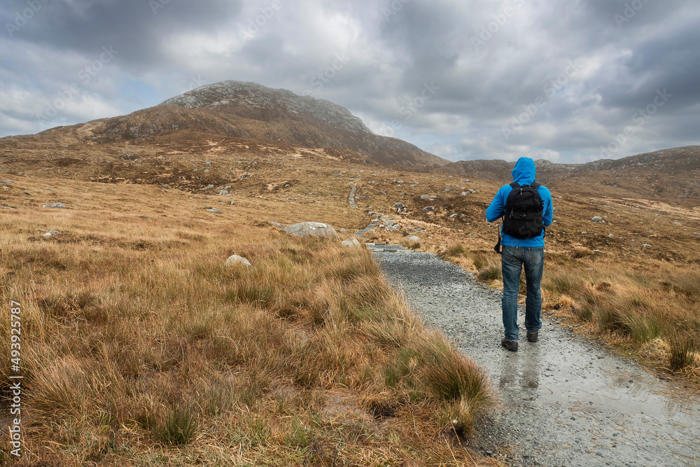 Male tourist walking towards Diamond hill, Connemara National Park, Ireland. Travel and hiking concept. Popular tourist area with stunning view. Irish nature landscape. Cloudy sky.