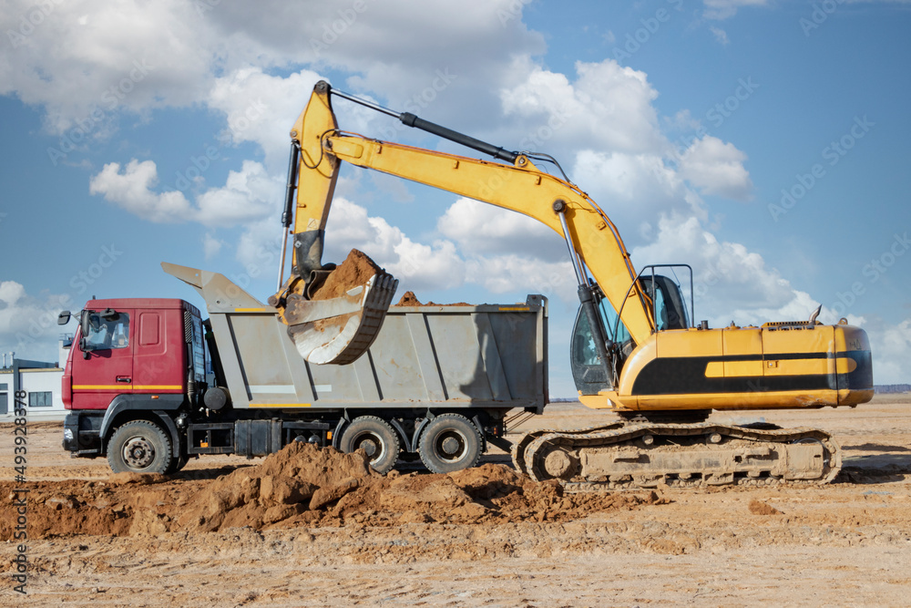 A powerful crawler excavator loads the earth into a dump truck against the blue sky. Development and removal of soil from the construction site. A powerful crawler excavator loads the earth into a dump truck against the blue sky. Development and removal of soil from the construction site.