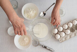 Process of making dough cake. woman’s hands mix flour and eggs, on the side stand milk and in bowl sugar on white table. ingredients for baking cake in kitchen, recipe. Top View.