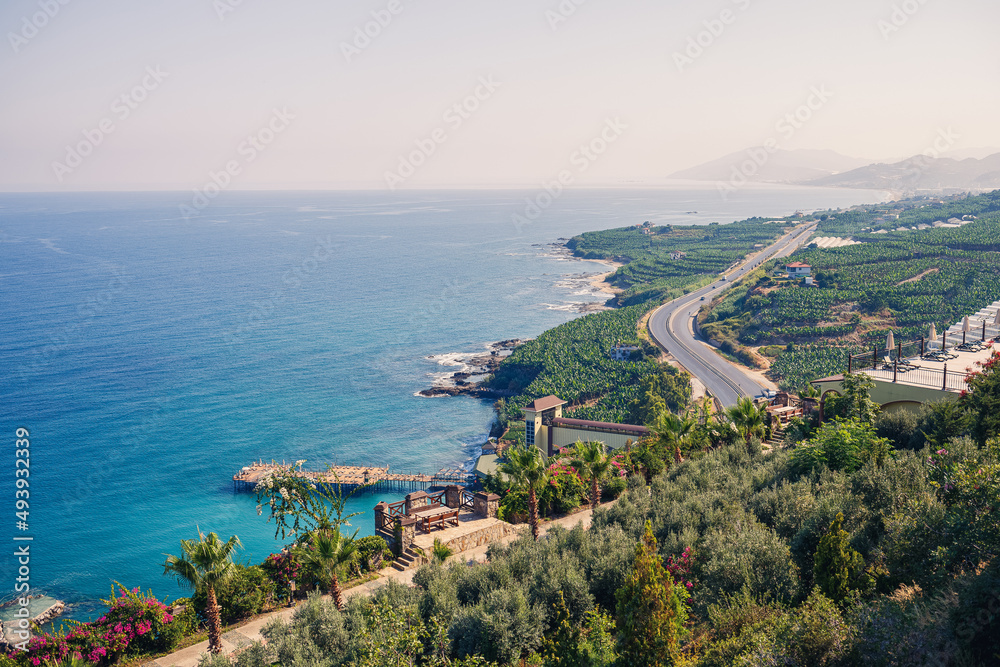 An empty asphalt road winds along the breathtaking scenic coastline on a sunny summer day. A spectacular shot of a coastal road overlooking clear blue skies and the calm Mediterranean Sea.