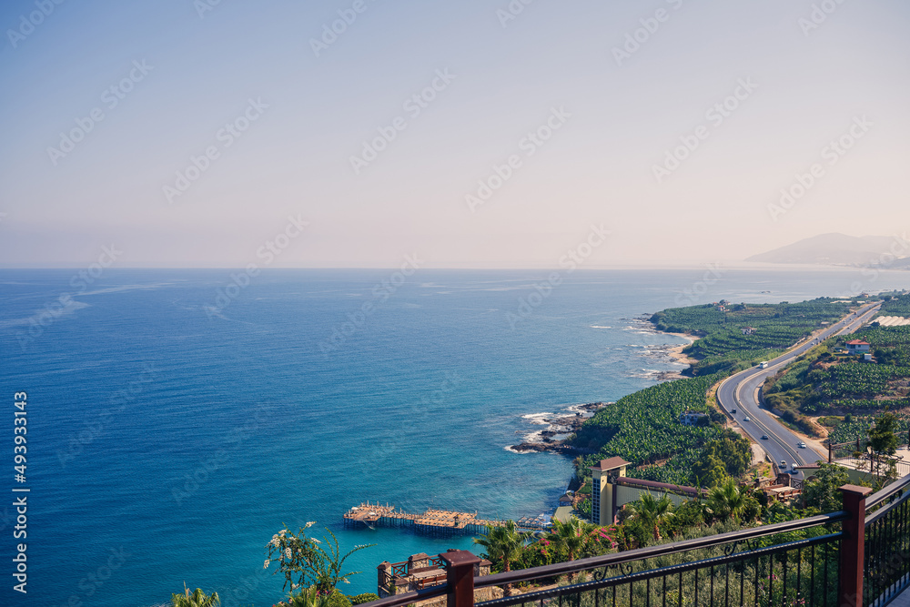 An empty asphalt road winds along the breathtaking scenic coastline on a sunny summer day. A spectacular shot of a coastal road overlooking clear blue skies and the calm Mediterranean Sea.